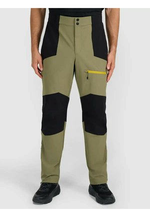 Mężczyzna noszący oliwkowo-zielone i czarne spodnie outdoorowe z żółtym zamkiem błyskawicznym na kieszeni na udzie oraz czarne buty sportowe.