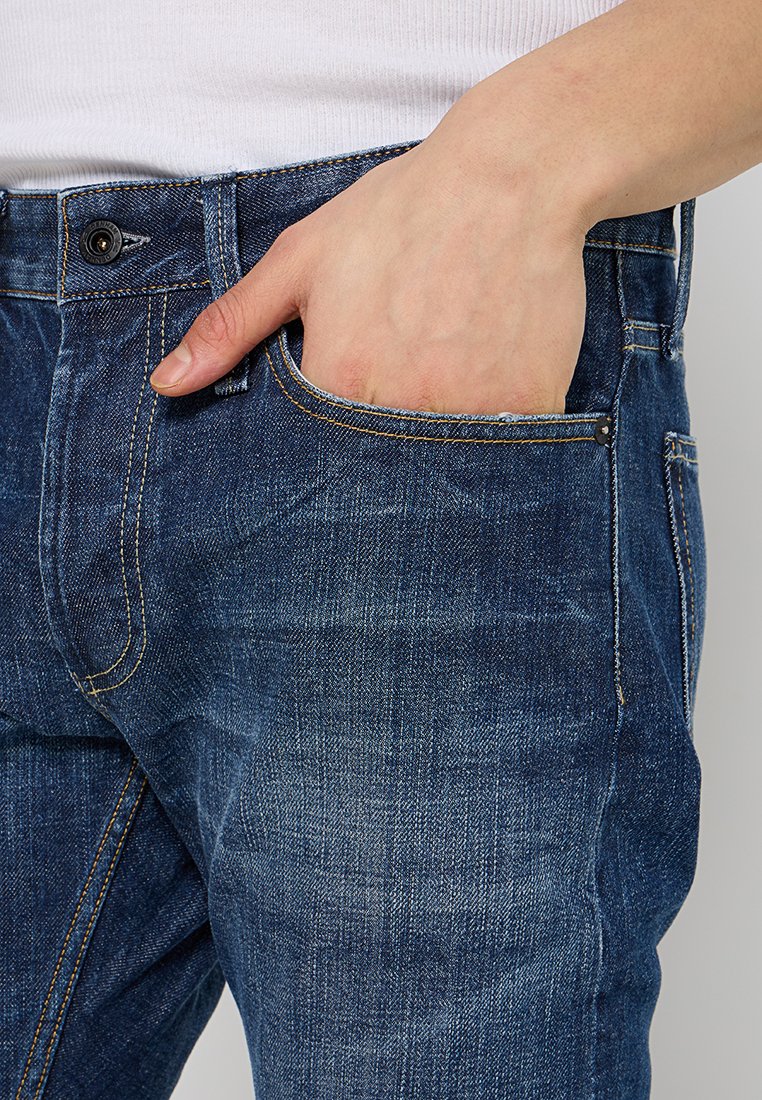 Donkerblauwe denim jeans met een vervaagde textuur, voorzien van gele stiksels, zijzak en een aansluitend ontwerp.