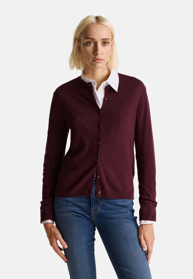 Burgunderfarbener Cardigan aus weichem Stoff, mit einer durchgehenden Knopfleiste, langen Ärmeln und gerippten Bündchen. Getragen über einem weißen Hemd mit Kragen.