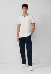Witte poloshirt met blauwe accenten, korte mouwen en een klein logo, gecombineerd met donkerblauwe corduroy broeken en witte sneakers.