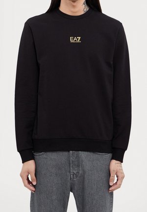 Zwarte lange mouwen sweatshirt met klein gouden "EA7 Emporio Armani" logo op de borst, gedragen met grijze jeans en zichtbare getatoeëerde handen.