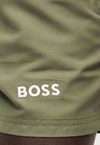 Λινό ύφασμα ελιάς με λευκό κεντημένο κείμενο "BOSS" κοντά στην κάτω άκρη, που δείχνει μέρος σκούρου δέρματος από κάτω.