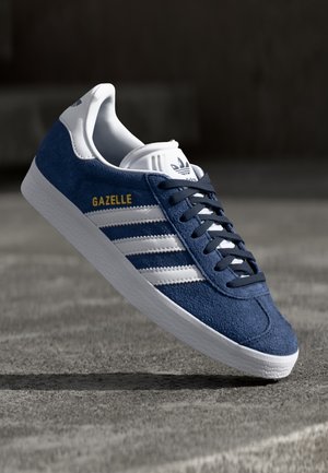 Blå Adidas Gazelle sneaker med hvide striber og sål, sorte snørebånd og guld "GAZELLE" tekst på ruskindsoverdel.