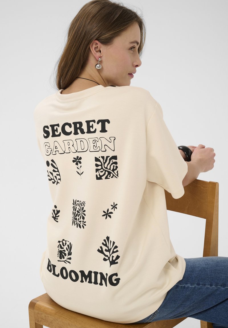 Jeune femme assise sur une chaise en bois, portant un t-shirt beige oversize avec texte noir "SECRET GARDEN BLOOMING" et des motifs floraux au dos.