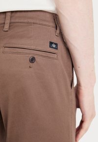 Brown cotton trousers with a back pocket, single button closure, and a small black logo patch. Smooth texture and tailored fit.