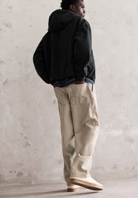 Fekete kapucnis dzseki, alatta kék csíkos ing, bézs kordbársony nadrág, krém színű sneaker barna talppal, egy texturált fal előtt áll.