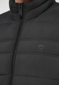 Chaqueta puffer negra con un cuello alto, textura acolchada horizontal y un logo negro en la zona izquierda del pecho. Cuenta con un cierre frontal.