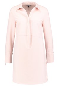 Jasnoróżowa tunika z kołnierzem, zapinana z przodu, z kieszenią na piersi, długimi rękawami oraz wiązaniem przy mankietach. Gładka faktura materiału.