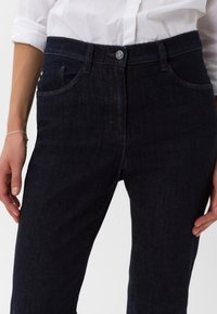 Tmavě modré džíny z denimu s přiléhavým střihem, s poutky na opasek, standardními kapsami a zapínáním na předním knoflíku. Hladká textura, minimální detaily šití.