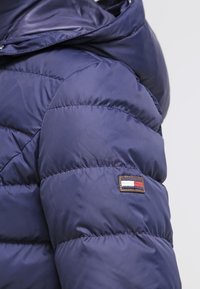 Námořnická modrá prošívaná bunda s kapucí, rukáv s malou obdélníkovou nášivkou s červenou, bílou a modrou vlajkou poblíž ramene.