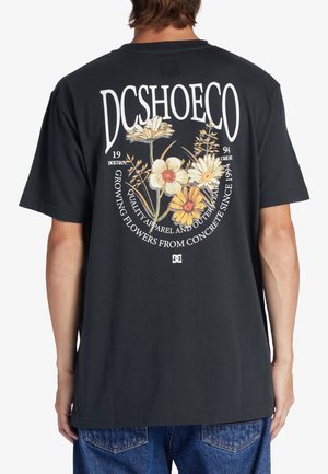 Osoba nosząca czarną koszulkę z kwiatowym motywem i tekstem "DCSHOECO" oraz "Growing flowers from concrete since 1994" na plecach.
