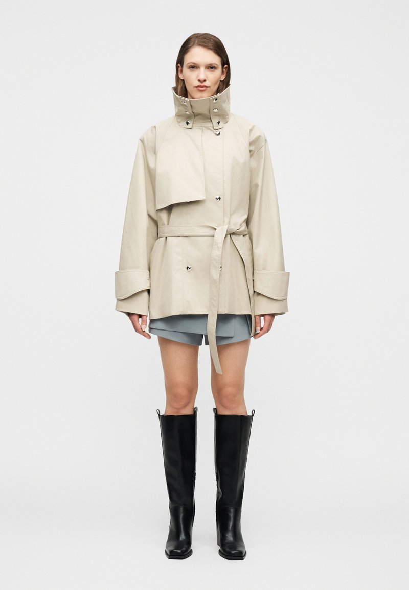 Γυναίκα που στέκεται φορώντας ένα μπεζ μπουφάν με ζώνη και ψηλό γιακά, γκρι φούστα και μαύρες μπότες μέχρι το γόνατο, σε λευκό φόντο.