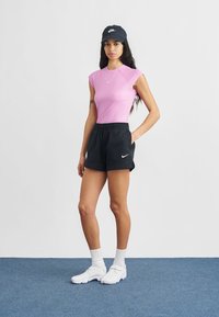 Růžový žebrovaný krátký rukáv, černé šortky s elastickým pasem, bílé logo Nike. Modelka má na sobě bílé ponožky a boty, černou čepici s logem.