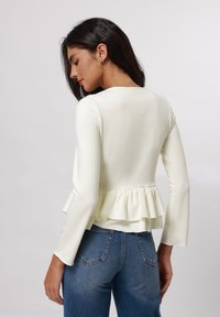 Top blanco de peplum con mangas largas, textura suave y dobladillo con volantes, combinado con jeans de mezclilla azul. La vista trasera muestra los detalles del diseño.
