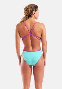 Donna con capelli lunghi, indossa un costume da bagno azzurro chiaro nella parte inferiore e un top viola con bretelle, in piedi con la schiena rivolta verso la parte anteriore su uno sfondo bianco.