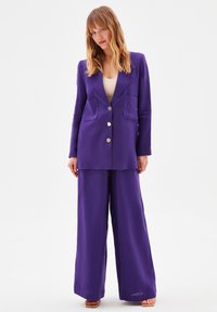 Completo in lino viola con blazer doppiopetto, due tasche e pantaloni a gamba larga. Texture liscia, design minimalista e bottoni in metallo.