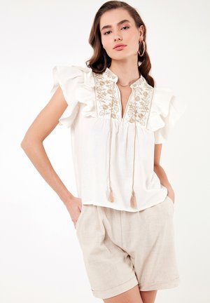 White blouse with ruffled sleeves and embroidered neckline, featuring a tassel tie detail. Paired with beige linen shorts.
