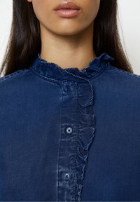 Navyblauwe blouse met een gerimpelde kraag en knoopsluiting. Het materiaal heeft een zachte textuur met een glanzende afwerking. De knopen zijn donkerblauw.
