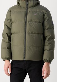 Olivgrön pufferjacka med huva, med en främre dragkedja, elastiska ärmmuddar och en liten logotyp på bröstet. Slät, quiltad yta.