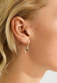 Goldene Creolen mit kleinen Charms in Grün, Weiß und Gold, die am linken Ohr einer blonden Person getragen werden, mit sichtbarer Hautstruktur und Muttermalen.