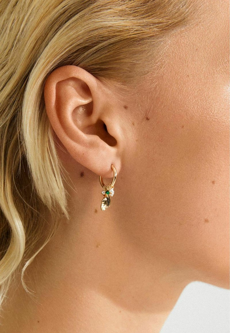 Goldene Creolen mit kleinen Charms in Grün, Weiß und Gold, die am linken Ohr einer blonden Person getragen werden, mit sichtbarer Hautstruktur und Muttermalen.