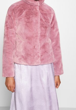 Femme portant une veste courte en fausse fourrure rose pâle sur une jupe texturée violet clair, debout devant un fond uni.