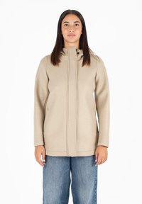 Cappotto beige con colletto alto, zip anteriore e tasche laterali, realizzato in tessuto morbido. Abbinato a jeans blu larghi.