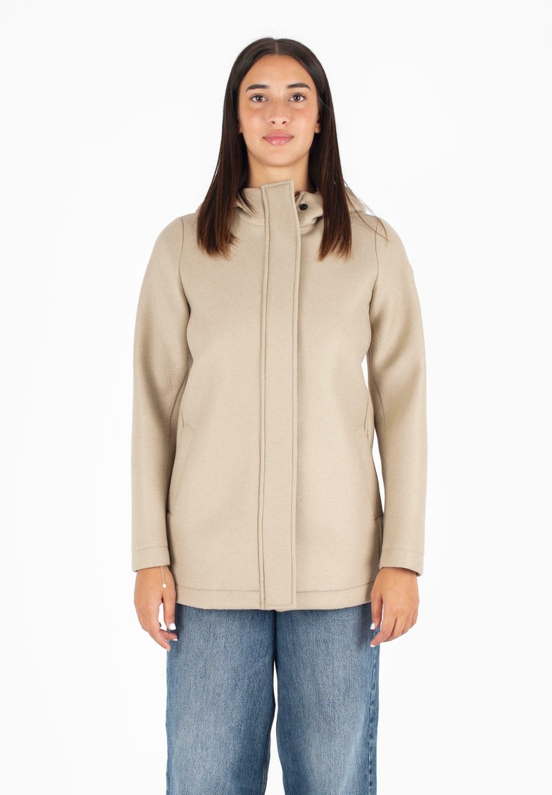 Cappotto beige con colletto alto, zip anteriore e tasche laterali, realizzato in tessuto morbido. Abbinato a jeans blu larghi.
