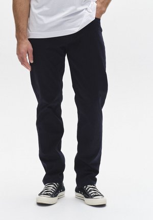 Mees, kes kannab musti pükse, must-valgeid tossusid ja valget särk, üks käsi taskus, valge taustaga.