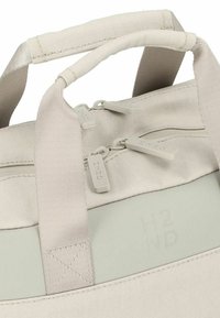 Sac en tissu gris clair avec deux poignées, fermeture éclair et logo "H2ND" en relief. Présente une texture lisse et un design minimaliste.
