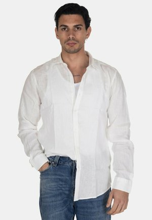 Uomo con capelli scuri che indossa una camicia di lino bianca con bottoni sopra una maglietta bianca e jeans blu, in piedi davanti a uno sfondo semplice.