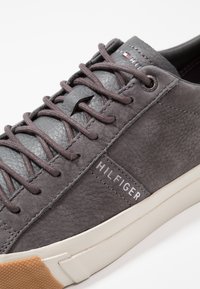 Detailní záběr na šedou tenisku Hilfiger s texturovanou kůží, odpovídajícími tkaničkami, kovovými očky a bílou podešví s hnědým texturovaným špičkovým krytem.