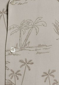 Camicia beige chiara con un motivo di palme marrone scuro, con chiusure a bottone sul davanti. Tessuto liscio, design semplice.