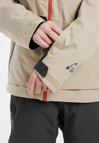 Beige waterdichte jas met een oranje rits, klittenband manchetten en zwarte accenten; heeft een ritszak op de mouw voor extra gemak.