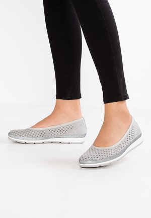 Fødder iført lysegrå perforerede slip-on sko med hvid sål og sølvglitterdetalje, kombineret med sorte tætsiddende bukser.