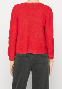 Czerwony, dziergany sweter z długimi rękawami, okrągłym dekoltem i krótkim krojem. Struktura swetra jest luźna i gruba. W komplecie z ciemnymi spodniami.