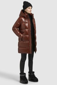Bruine puffer jas met een glanzende afwerking, ritsluiting en capuchon, gecombineerd met zwarte leggings en stevige zwarte laarzen.