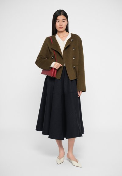 Olive rohelise topeltk inglise villane jakk suurte nuppudega, sobitatud kreemja kampsuni ning musta volditud maxi seelikuga. Mudel kannab valgeid mulu.