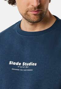 Felpa blu navy realizzata in morbido tessuto, con scollatura rotonda e testo stampato bianco: "Slade Studios Original Wear" e "DESIGNED FOR YOUR FREEDOM."