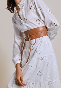 Witte blouse met geborduurde bloemen uitsparingen, voorzien van een tan leren riem. De blouse heeft een knoopsluiting kraag en wijd uitlopende mouwen.