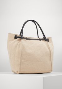 Beige tygväska gjord av canvas med mörka läderremmar och sömmar. Väskan har en rymlig silhuett och en minimalistisk design.