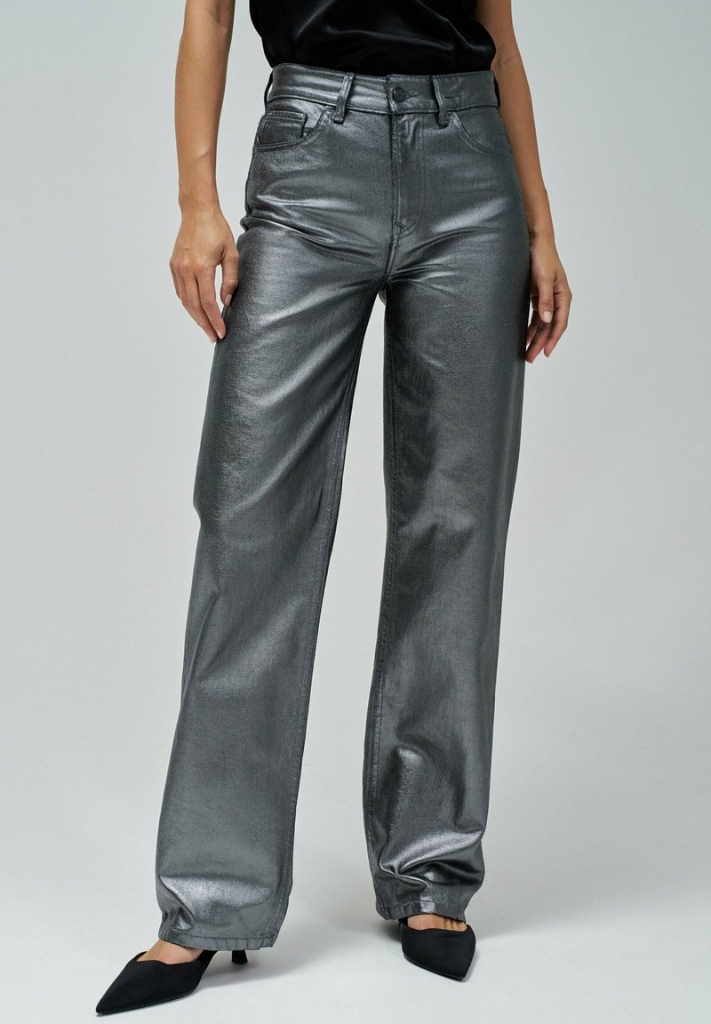 Pantalons large en gris métallique avec une texture lisse, taille haute, fermeture à bouton et ourlets retroussés, associés à des chaussures noires à bout pointu.