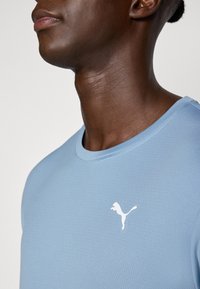 Chemise à manches courtes texturée bleu clair. Design à col rond avec un logo blanc sur la poitrine. Le tissu semble lisse et respirant.