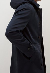 Laivastonsininen takki hupulla, jossa on napillinen etuosa, sivutaskut ja sileä pinta. Räätälöity malli suoralla siluetilla.