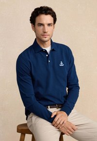 Man met donker krullend haar, gekleed in een blauwe lange mouwen polo en beige broek, zittend op een houten kruk tegen een neutrale achtergrond.