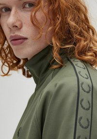 Giacca zip-up verde oliva con strisce laterali decorate e colletto alto. Presenta un tessuto testurizzato e un logo ricamato sul petto.