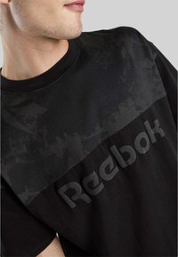 Joven con una camiseta negra de Reebok que presenta un sutil patrón texturizado en el pecho y un logo en relieve.
