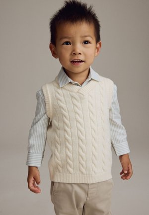 Ung dreng med kort pigget hår iført en hvid kabelstrikket ærmeløs sweater over en stribet skjorte og beige bukser, stående mod en ensfarvet baggrund.