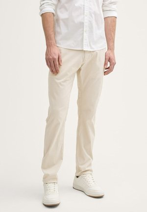 Uomo con pantaloni color crema, scarpe da ginnastica bianche e camicia bianca a maniche lunghe con maniche rimboccate, in piedi su uno sfondo chiaro.