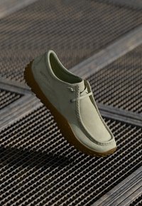 Zapato de ante verde con punta redondeada, suela de goma marrón con un patrón texturizado y detalles de costura en contraste. Un diseño minimalista de cordones.